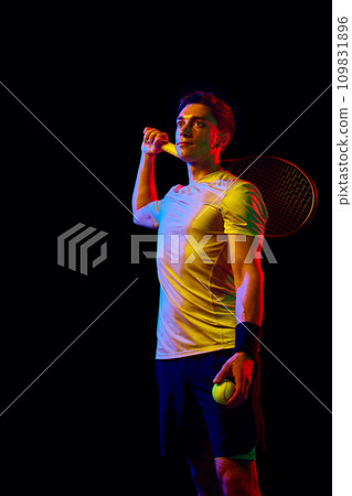 Portrait of athletic man, tennis player standing, posing with racket and ball against dark background in neon light 109831896