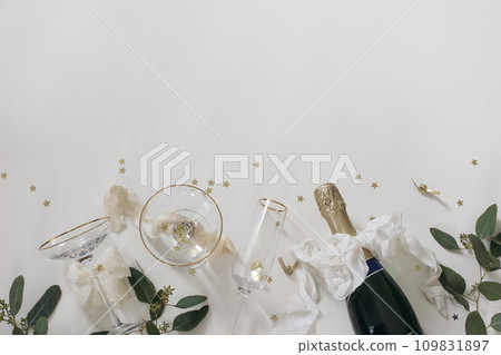 New year party, celebration banner. Bottle of champagne wine, drinking glasses, bows. Eucalyptus tree branches isolated on white table background. Golden star confetti. Birthday flat lay, top view. 109831897