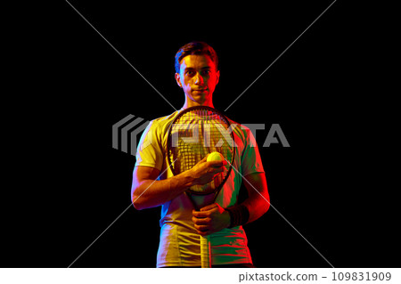 Portrait of athletic man, tennis player standing, posing with racket and ball against dark background in neon light 109831909