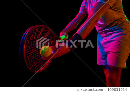 Cropped image of male tennis player in blue shirt and white shorts, serving ball with racket against dark background in neon light 109831914