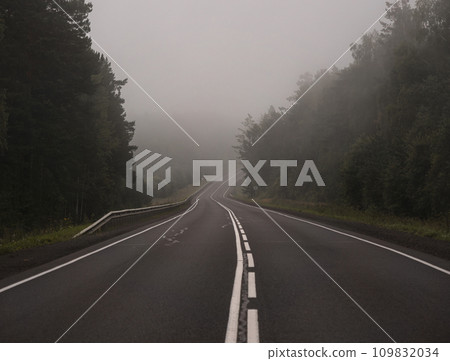 Empty asphalt road in foggy forest. Curved roadway, trees with green foliage in fog and overcast sky. Landscape with empty asphalt road through woodland in summer. Empty asphalt road in foggy forest. Curved roadway, trees with green foliage in fog and overcast sky. Landscape with empty asphalt road through woodland in summer. 109832034