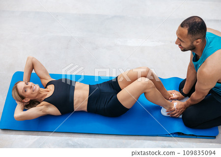 Woman working on sit-ups while personal trainer provides assistance. Woman working on sit-ups while personal trainer provides assistance. 109835094