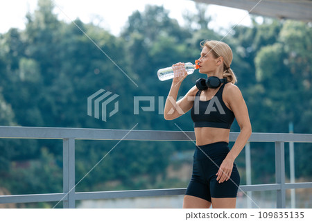Woman with headphones resting and drinking water after running. Woman with headphones resting and drinking water after running. 109835135
