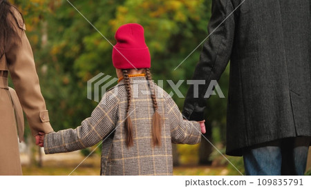 Loving little girl joins hands with mother and father walking across autumn garden. Little daughter enjoys weekend with parents in park. Family of mother, father and little daughter in fall garden Loving little girl joins hands with mother and father walking across autumn garden. Little daughter enjoys weekend with parents in park. Family of mother, father and little daughter in fall garden 109835791