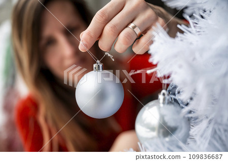Young woman placing ice blue holiday bauble on a white christmas tree 109836687