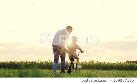Gentle father supports toddler girl training bicycle riding in sunset field 109837130