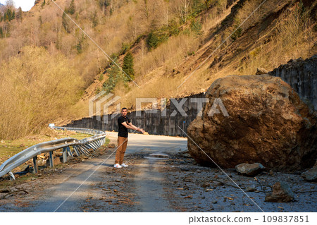 A huge rock fell from the mountains onto the road, destroying the asphalt and blocking half of the roadway 109837851