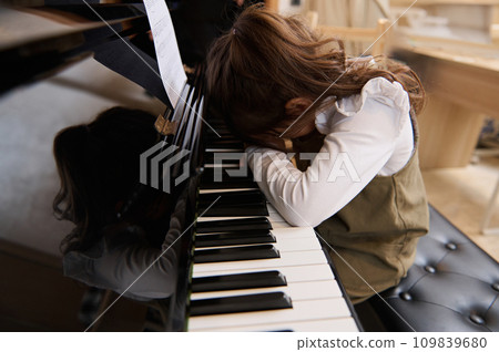 Cute little girl folds her hands and hid her face, feeling sadness, having difficulties or forced to learn playing piano 109839680