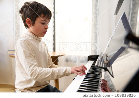 Adolescent boy performing classical composition, touching the keys and feeling rhythm while playing grand piano at home Adolescent boy performing classical composition, touching the keys and feeling rhythm while playing grand piano at home 109839826
