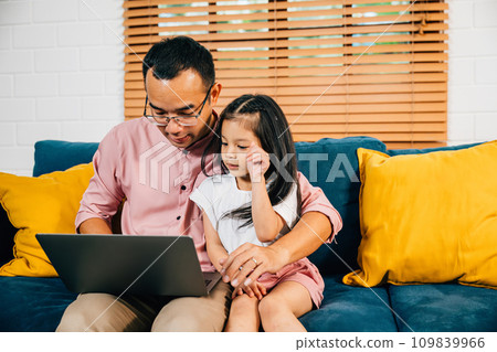 An Asian father finds happiness in his work on a laptop while his daughter enjoys e-learning on a computer in their modern living room. Their togetherness and smiles create a special family moment. 109839966