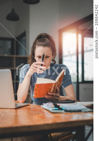 Business woman sitting at office desk and writing notes in a notebook while talking on a video call meeting with clients or coworkers on her laptop computer. Business woman sitting at office desk and writing notes in a notebook while talking on a video call meeting with clients or coworkers on her laptop computer. 109842242
