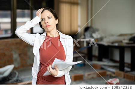 Confused female designer standing with papers in apartment during renovation 109842338