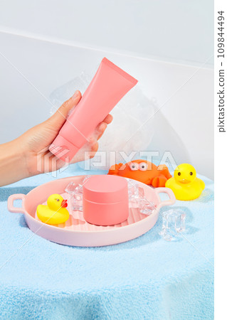 A hand holding a tube of pink cosmetics, a jar of lotion, silicone bath toys, a bath sponge, ceramic tray and ice are placed on a table covered with a blue towel. Skin care concept. A hand holding a tube of pink cosmetics, a jar of lotion, silicone bath toys, a bath sponge, ceramic tray and ice are placed on a table covered with a blue towel. Skin care concept. 109844494