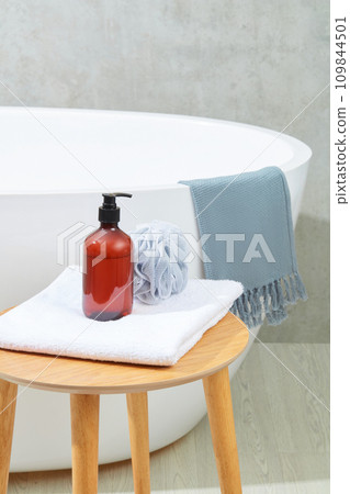 A bottle of brown shampoo and a bath sponge on a folded towel on a wooden table, next to a bathtub on a gray background. Space inside the bathroom for advertising. A bottle of brown shampoo and a bath sponge on a folded towel on a wooden table, next to a bathtub on a gray background. Space inside the bathroom for advertising. 109844501
