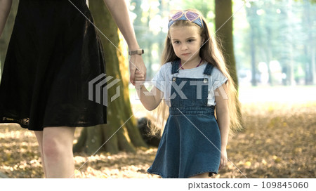 Mom and her small daughter with long hair walking together holding hands in summer park. 109845060