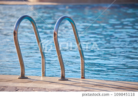 Close up of swimming pool stainless steel handrail descending into tortoise clear pool water. Accessibility of recreational activities concept. 109845118