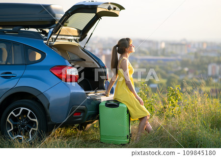 Young woman resting on a green suitcase near her car in summer nature. Travel and vacations concept. 109845180