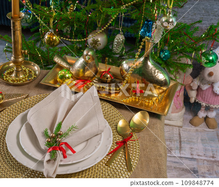 New Year's table setting in gold color, against the background of a decorated Christmas tree. 109848774
