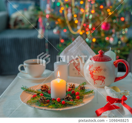 On the table with a linen napkin is a burning candle, a mug of tea, a teapot against the backdrop of a Christmas tree 109848825