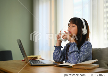 Asian schooler girl drinking milk while doing homework or studying online on laptop at home. 109854072