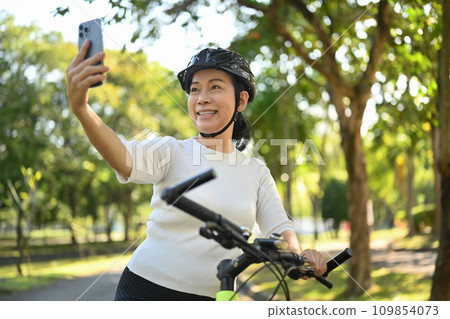 Happy middle age woman sitting on a bicycle and making a video call on mobile phone 109854073
