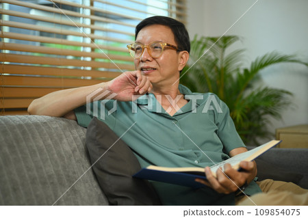 Happy middle age man resting on comfortable sofa holding book and looking away. 109854075