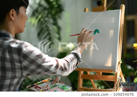 Side view of young man painting on canvas in garden. Art and leisure activity concept. 109854076