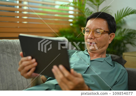 Carefree senior man browsing internet on digital tablet while relaxing on a couch at home. 109854077
