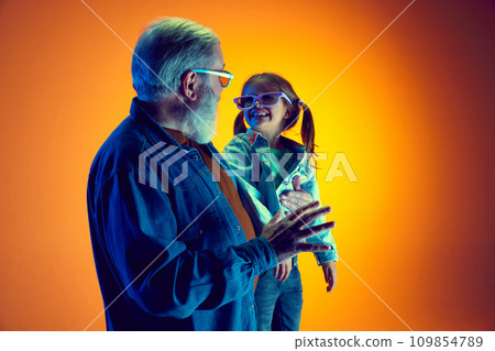Senior man and little girl, grandfather and granddaughter watching TV over gradient orange background in neon light Senior man and little girl, grandfather and granddaughter watching TV over gradient orange background in neon light 109854789