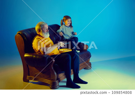 Senior man and little girl, grandfather watching TV with his granddaughter over blue background in neon light Senior man and little girl, grandfather watching TV with his granddaughter over blue background in neon light 109854808