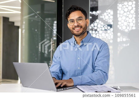 Portrait of young successful businessman inside office, satisfied joyful Indian man looking at camera, worker at workplace using laptop, typing on keyboard, financier accountant in shirt. 109855561