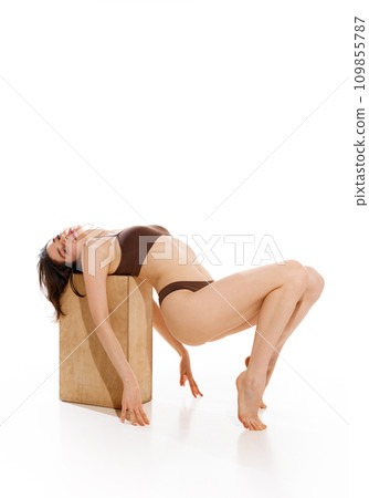 Creative full length portrait of slender young woman in brown underwear posing on wood cube against white studio background. Creative full length portrait of slender young woman in brown underwear posing on wood cube against white studio background. 109855787