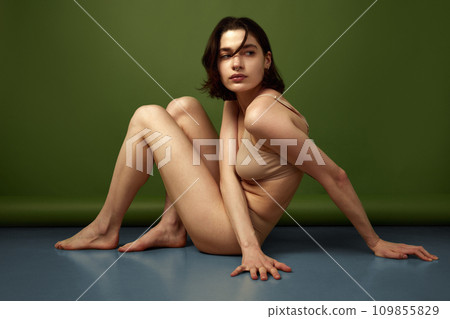 Side view portrait of brunette woman posing sitting on floor in flesh color lingerie and looking away against olive studio background. Side view portrait of brunette woman posing sitting on floor in flesh color lingerie and looking away against olive studio background. 109855829