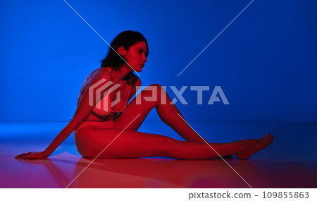 Essence of contemporary beauty. Beautiful, young woman posing on floor in lingerie and looking away against gradient background in neon. Essence of contemporary beauty. Beautiful, young woman posing on floor in lingerie and looking away against gradient background in neon. 109855863