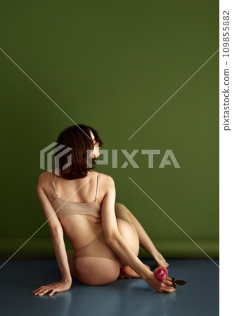 Rear view portrait of attractive slender lady, posing holding red blooming flower sitting on floor and wearing flesh color lingerie against olive background. Rear view portrait of attractive slender lady, posing holding red blooming flower sitting on floor and wearing flesh color lingerie against olive background. 109855882