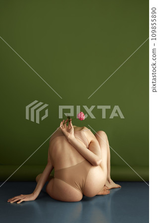 Young, beautiful woman sits with her back to camera and poses holding red blooming flower behind her back against olive studio background. 109855890