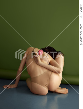 Rear view of slim female body. Woman with healthy back, sitting on floor and posing holding red flower against olive studio background. Rear view of slim female body. Woman with healthy back, sitting on floor and posing holding red flower against olive studio background. 109855895