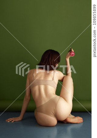 Rear view of fit female body. Woman with healthy back, sitting on floor and posing holding red flower against olive studio background. 109855899