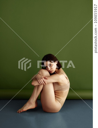 Naked, brunette woman posing dressed beige lingerie sitting hugging legs on floor and looking at camera against olive studio background. 109855917