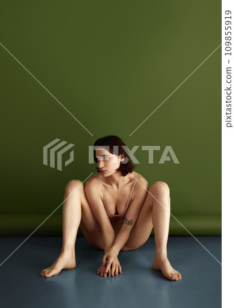Naked, brunette woman, flexible model posing dressed beige lingerie sitting on floor against olive studio background. Naked, brunette woman, flexible model posing dressed beige lingerie sitting on floor against olive studio background. 109855919