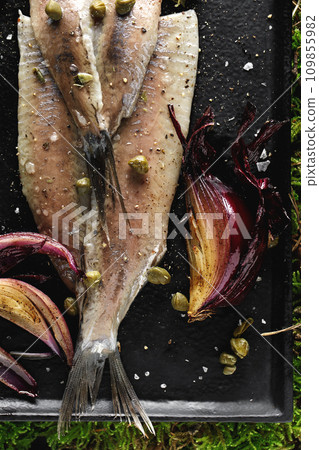Herring fillet with capers, salt flakes, red onion and pepper served on black tray on green forest moss background. Food aesthetic concept 109855982