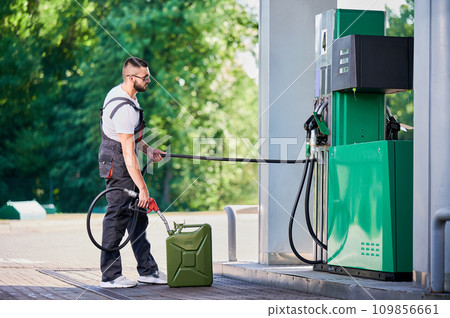 Professional worker refueling canister with fuel. Skilled man filling cistern with gasoline for customer. Man in ovealls with pump nozzle refueling canister next to gasoline column. Professional worker refueling canister with fuel. Skilled man filling cistern with gasoline for customer. Man in ovealls with pump nozzle refueling canister next to gasoline column. 109856661