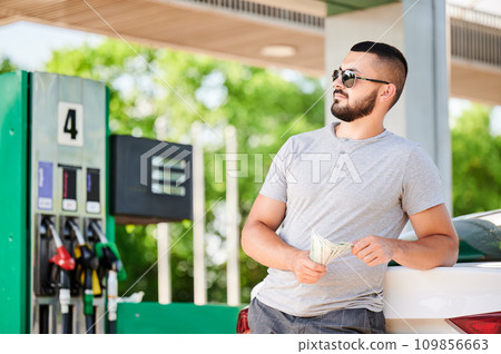 Young man waiting for petrol station operator to pay for fuel. Man leaning on his car and counting how much gasoline costs. Male in casual clothes holding money and looking away. Young man waiting for petrol station operator to pay for fuel. Man leaning on his car and counting how much gasoline costs. Male in casual clothes holding money and looking away. 109856663
