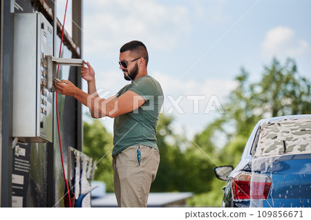 Young man paying for cleaning car on self-service car wash. Cropped image of blue auto and man that going to wash it by active foam. Male person paying for wash service next to car at sunny day. Young man paying for cleaning car on self-service car wash. Cropped image of blue auto and man that going to wash it by active foam. Male person paying for wash service next to car at sunny day. 109856671