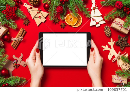 Top view of woman holding tablet in her hands on red background made of Christmas decorations. New Year holiday concept. Mockup 109857701