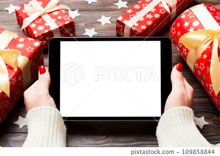 Top view of female hands holding a tablet on wooden Christmas background made of gift boxes and snowflakes. New year holiday concept. Mockup Top view of female hands holding a tablet on wooden Christmas background made of gift boxes and snowflakes. New year holiday concept. Mockup 109858844