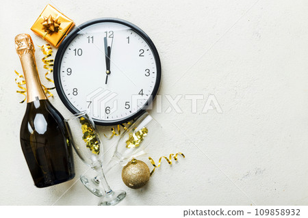 New year celebration concept with a bottle of champagne and two glasses toasting. Christmas gift box, alarm clock and fir tree branch on colored table. Top view Copy space 109858932