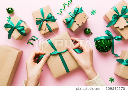 Woman is packing surprise gift for Christmas holidays, sitting at workplace with craft tools, top view. Woman wrapping Christmas gift at colored table Woman is packing surprise gift for Christmas holidays, sitting at workplace with craft tools, top view. Woman wrapping Christmas gift at colored table 109859270