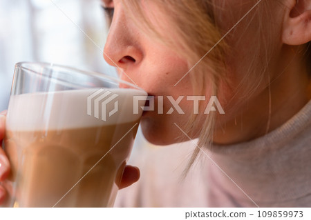 Woman with blonde hair sips cappuccino in a cafe. She is holding the glass up to her face, taking a sip of the drink. 109859973