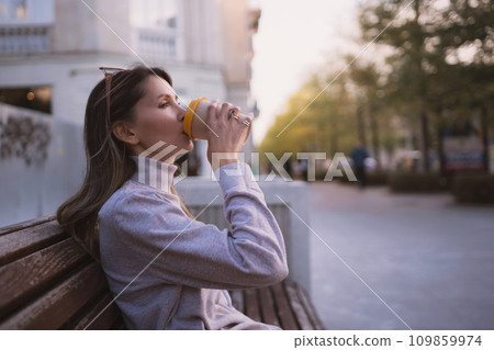 Woman drinks from cup on wooden bench. She is wearing a white shirt enjoying her beverage. The bench is located in a park setting, with trees in the background. 109859974